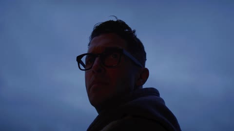 A man wearing glasses and a coat is standing outside and as he turns around and looks ahead a shadow moves over him
