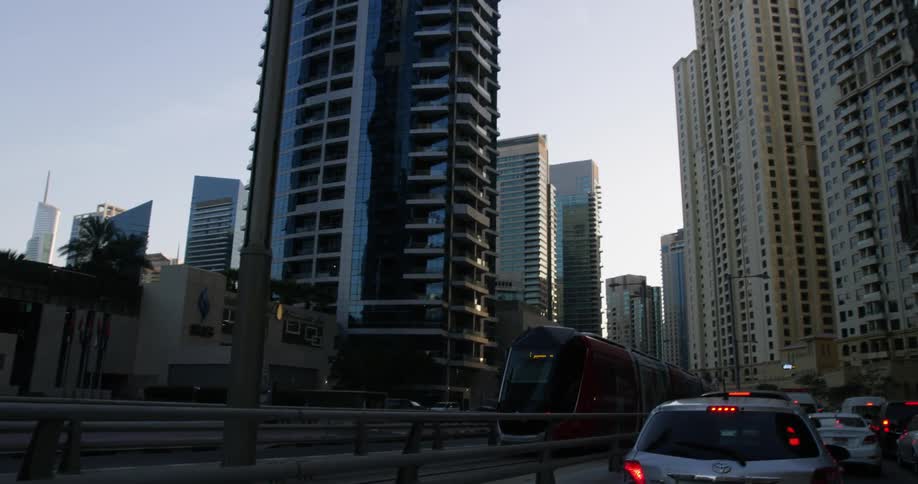 A train passes through downtown surrounded by tall buildings and skyscrapers as traffic is stopped on the street