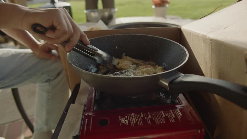 People hang around and watch as person flips food frying in skillet on portable burner