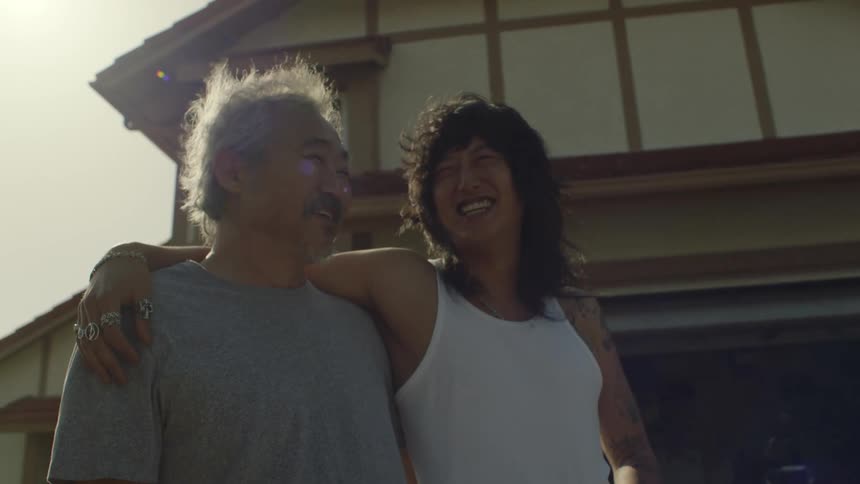 Two asian men are outside in front of a house smiling and laughing as the sun shines brightly in the distance