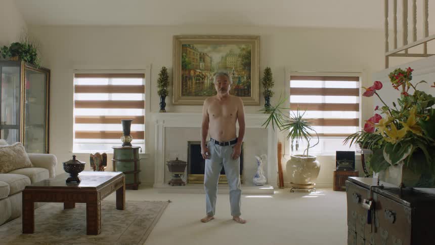A shirtless elderly asian man looks into the camera while standing in the living room of a house during the day