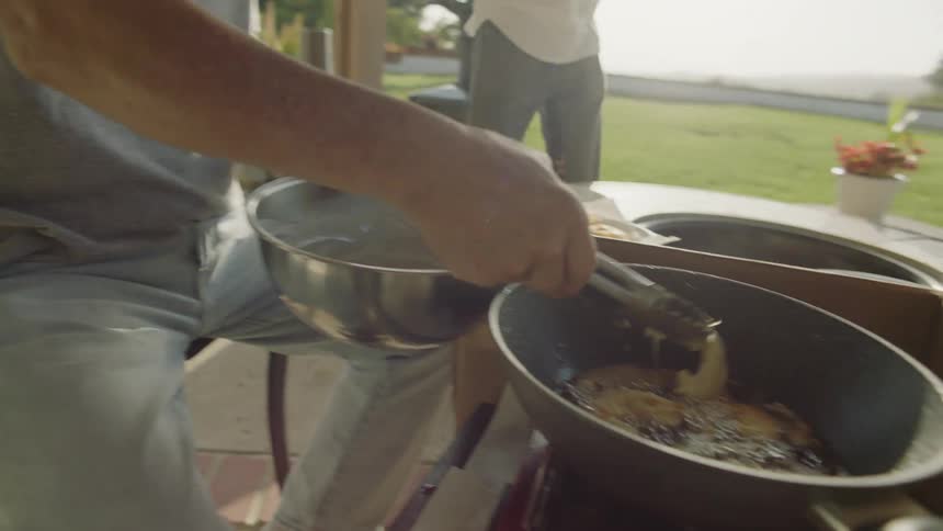 A man uses tongs to place food into a hot skillet while cooking outdoors during the day