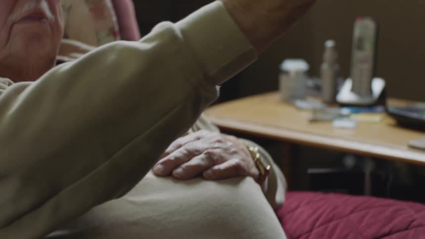 Elderly man sitting in chair reaches up to clasp hands with another person