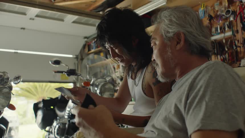 A father and son sit in a garage with motorcycles and look through photographs