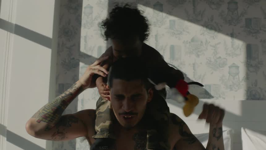 Dad with tattoos on chest arms and neck lifts his young son and puts him on his shoulders in a sun filled bedroom