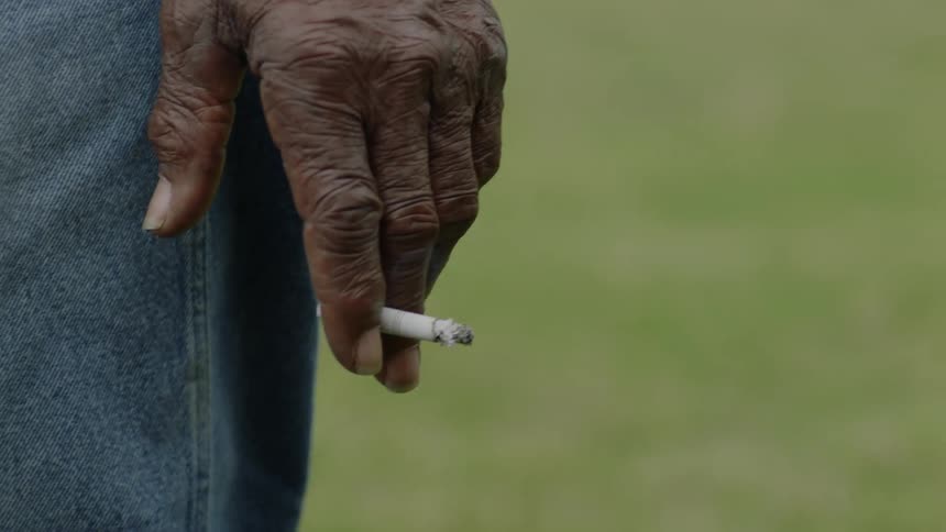 A man standing with one hand hooked onto his belt flicks his cigarette ash with the other
