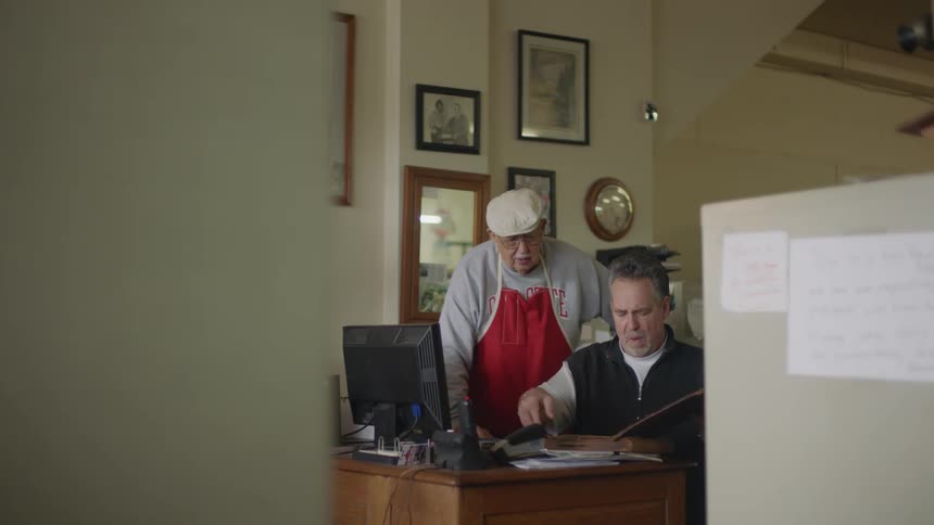 A man is sitting at a desk looking at a book and an older man wearing a red apron is standing next to him