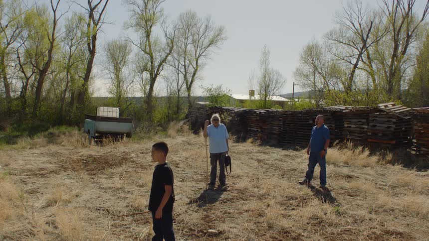 Three people are standing outside on dry grass near a fence and there is a building in the distance