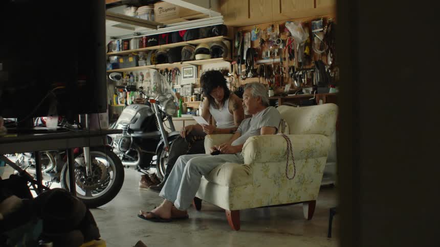 Two men sit in a garage by motorcycles and talk