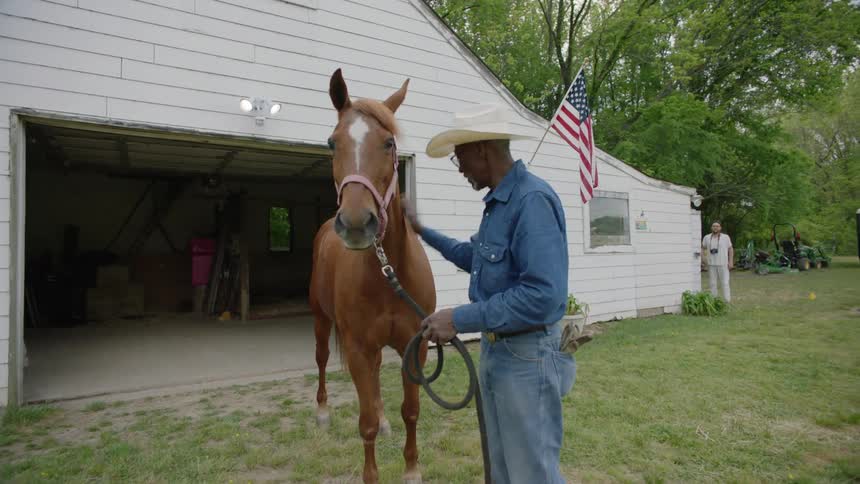 A man wearing a cowboy hat stands next to a colt petting him and talking