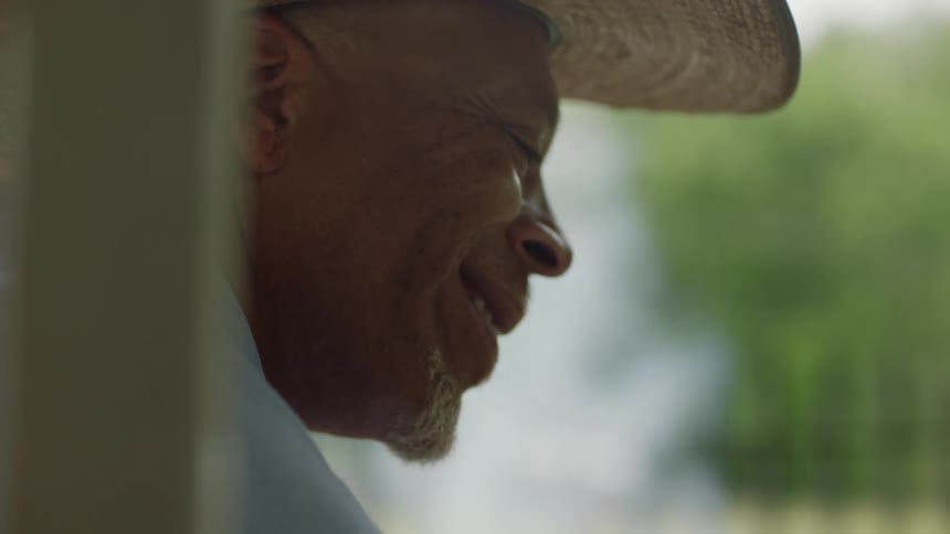 A man wearing a cowboy hat is outside speaking to someone and there is smoke in the distance