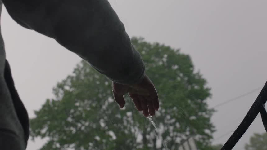 A man is standing on a porch with his hand out in the rainstorm letting water run from his fingers