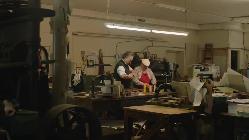 A man twists the top on a bottle and reaches to apply it for an elderly man in a workshop