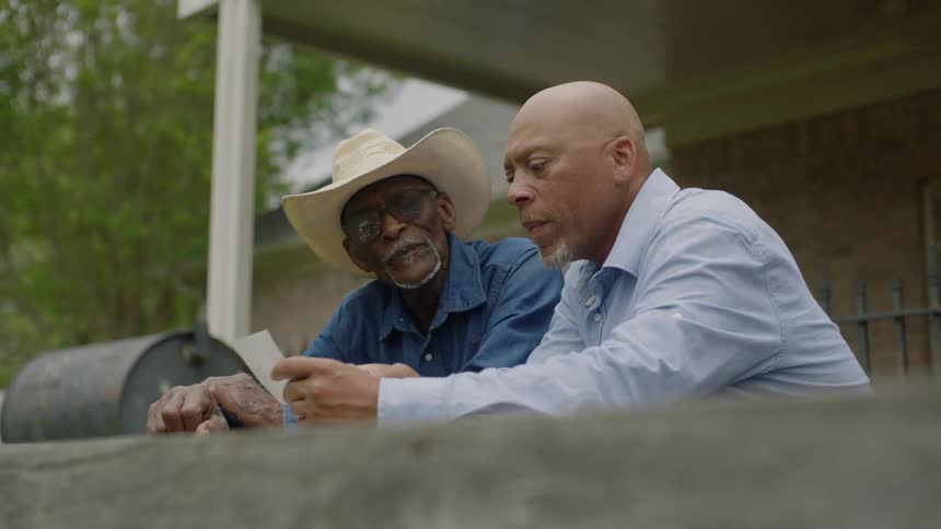Two older men are sitting on a porch looking at a paper