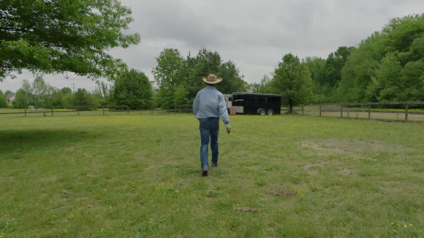 Male cowboy walks across pasture toward parked trailers