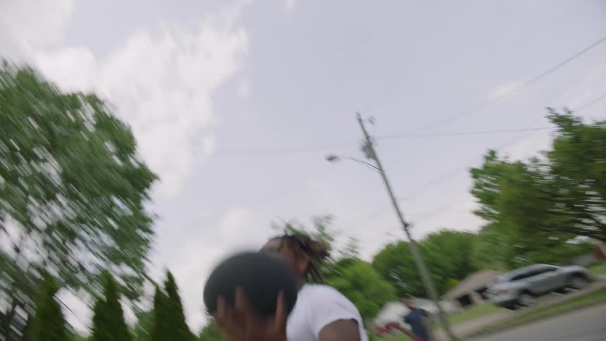 A teenage boy dribbles a basketball up a driveway in a neighborhood and shoots the ball into a hoop as his brother stands and watches