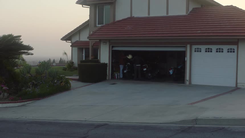 House garage door is raised and two men put on motorcycle helmets while standing near motorcycles