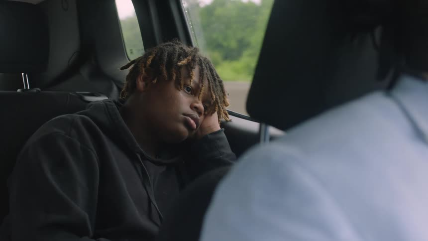 A teenager sits with his head on his hand in the back seat of a car