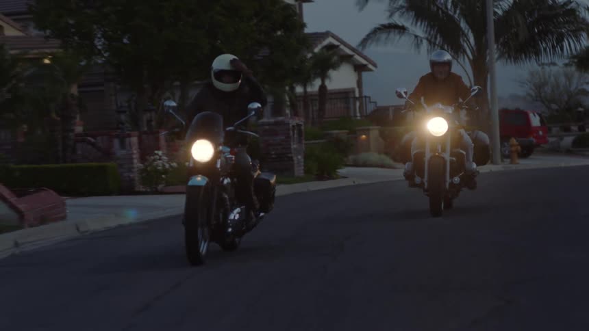 Two men ride motorcycles through a neighborhood in the evening