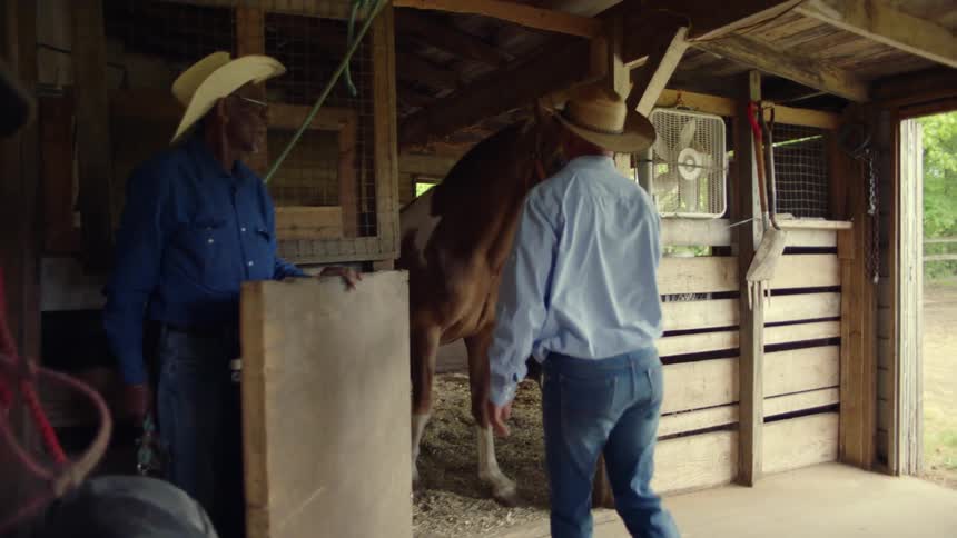 Two cowboys are in a barn and one opens a stall and the other walks out with a horse