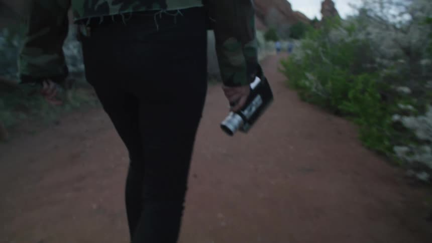 Woman walks along dirt path near mountain stopping to aim her video camera