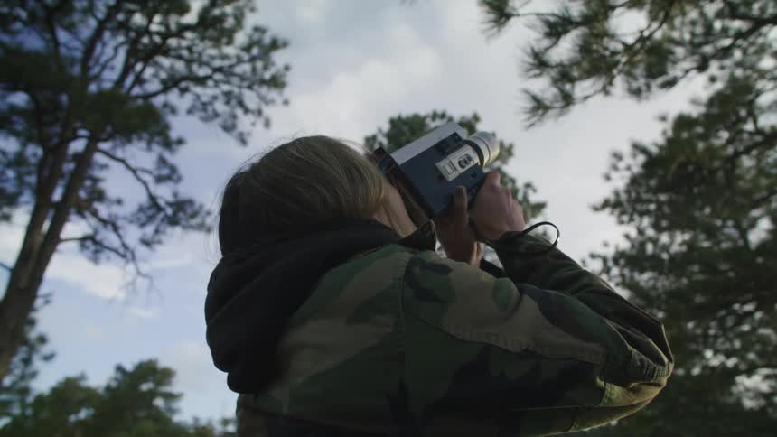 A woman is standing in a forest using a video camera to capture film