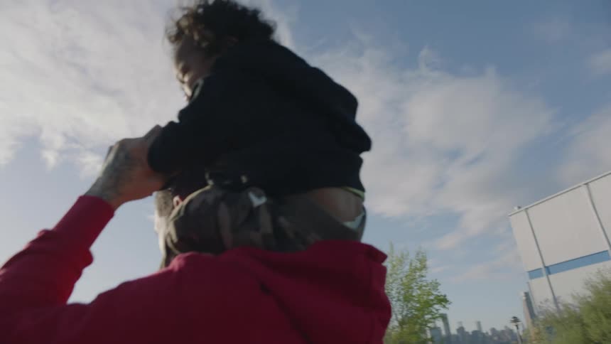 A man is outside at a park near water playing with his young son on his shoulders