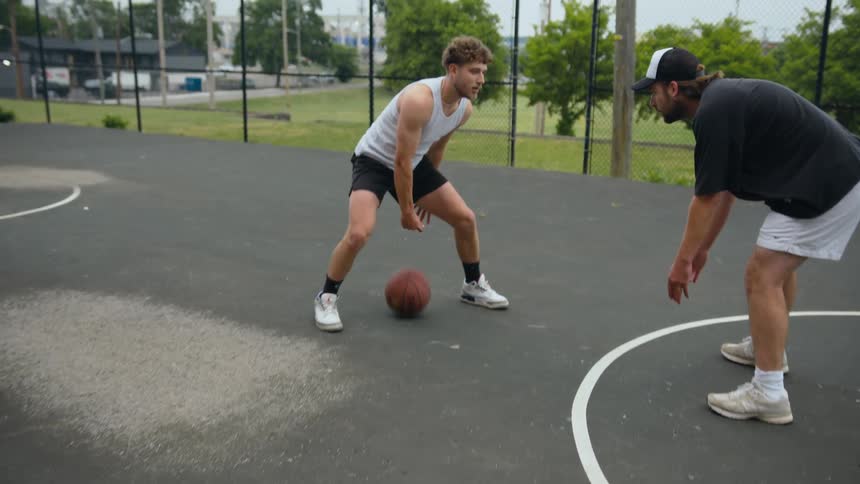 Two men play one on one basketball on court in park