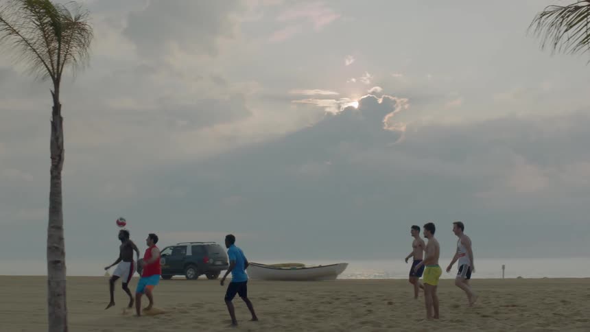A ground of guys are playing soccer on the beach and there is a truck driving by in the background