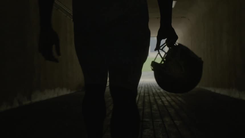Football player walks in a dark tunnel while carrying a football helmet