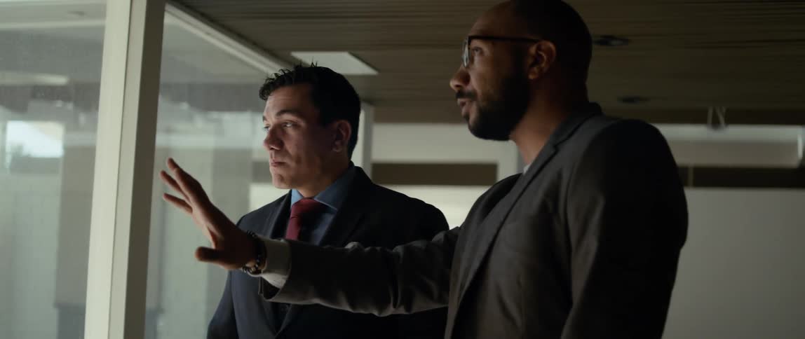 Two men are standing near a window and one man is using his hands as he talks