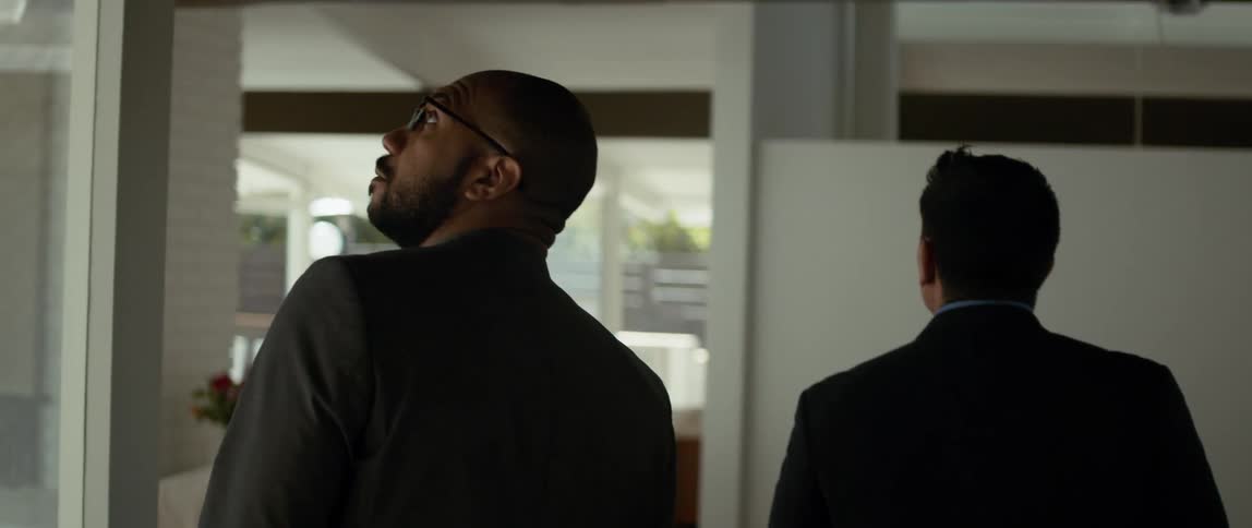 Two men are walking into a room and one man is looking around as the other man speaks