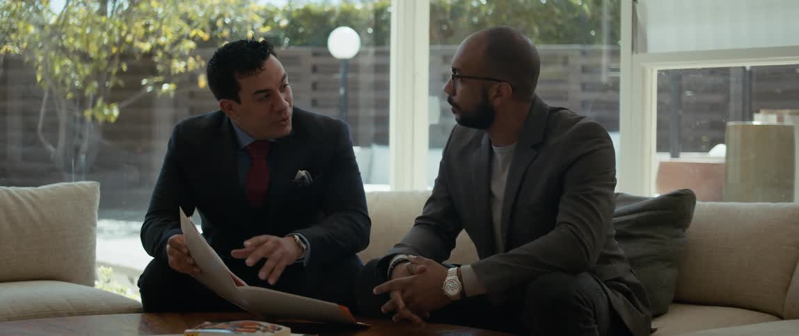 Two men are sitting on a couch looking at a notebook that one man is holding as he is speaking