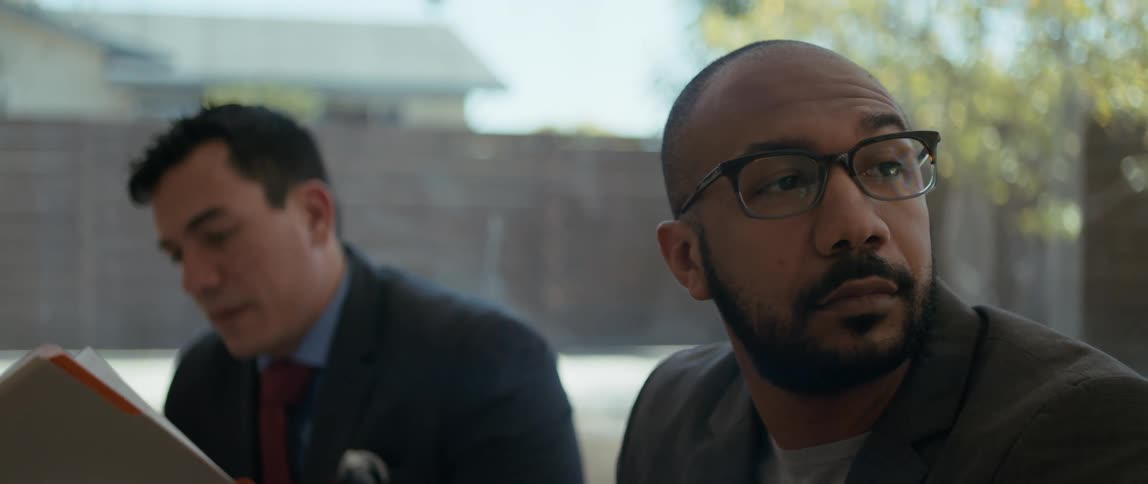 One man sits in an office and looks around as another man looks over papers