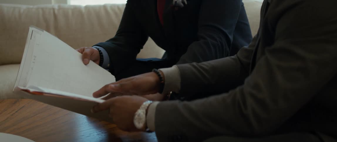 Two men discuss a folder of paperwork while sitting on a patio outdoors