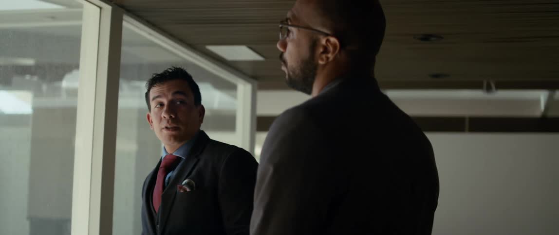 Two men stand in an office and talk as they look out a window