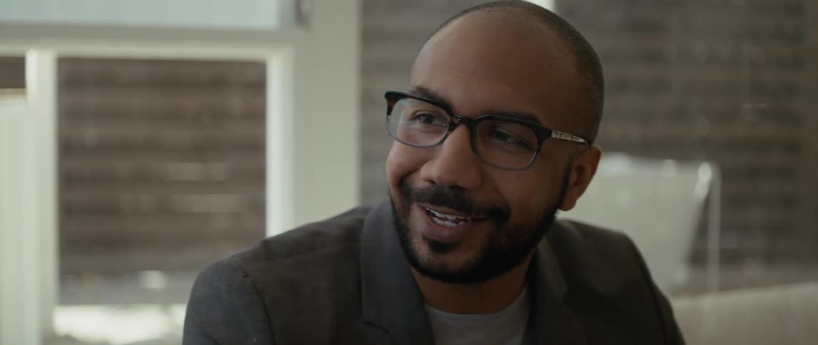 A man wearing glasses smiles as he speaks to someone