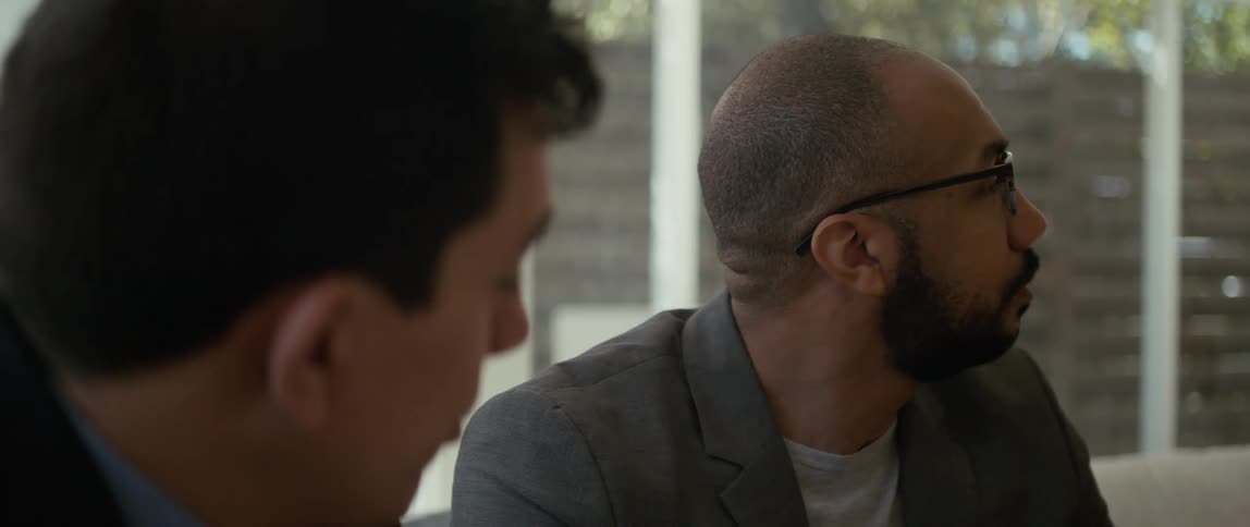 Two men are sitting together and one man is looking around as the other man speaks to him