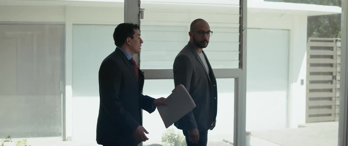 A realtor holding a folder talks with a client as they walk toward glass doors leading outside