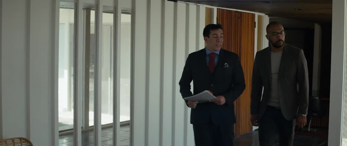 Two men are walking through a room as they talk and one man is carrying a notebook