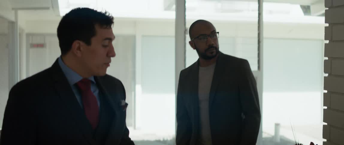 Two men are walking through a room as they talk and one man is carrying a folder