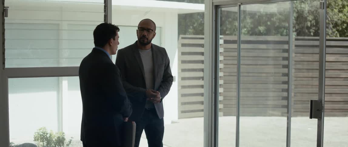 A realtor discusses a home with a client while they stand near the glass doors looking out onto a driveway