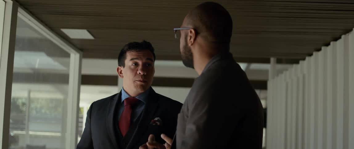 Two men are standing near a window and one man is speaking as the other man listens