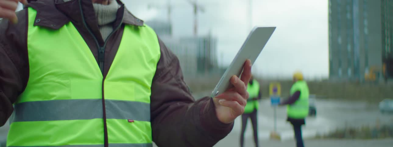 Man in a safety vest holds a tablet and talks to it as two people stand behind him
