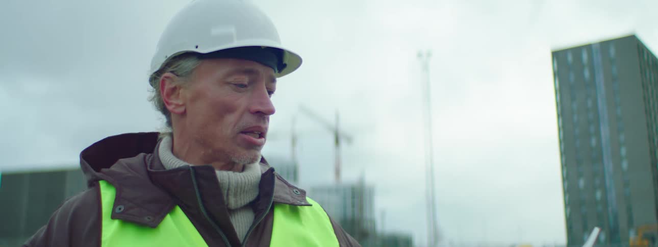 Man in a hard hat and safety vest talks and motions with his hand at a construction site
