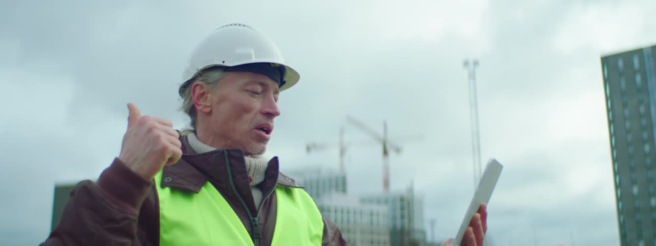 Construction worker stands at work site surrounded by buildings talking to someone via tablet