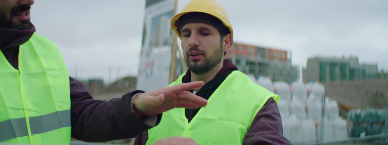 Two male construction workers wearing safety helmets and green vests standing in a construction site and talking about the ongoing construction