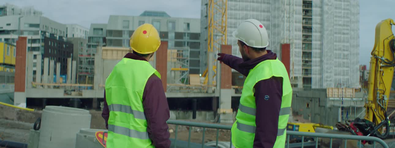 Two male construction workers at a construction site wearing safety helmets and green vests talking to each other