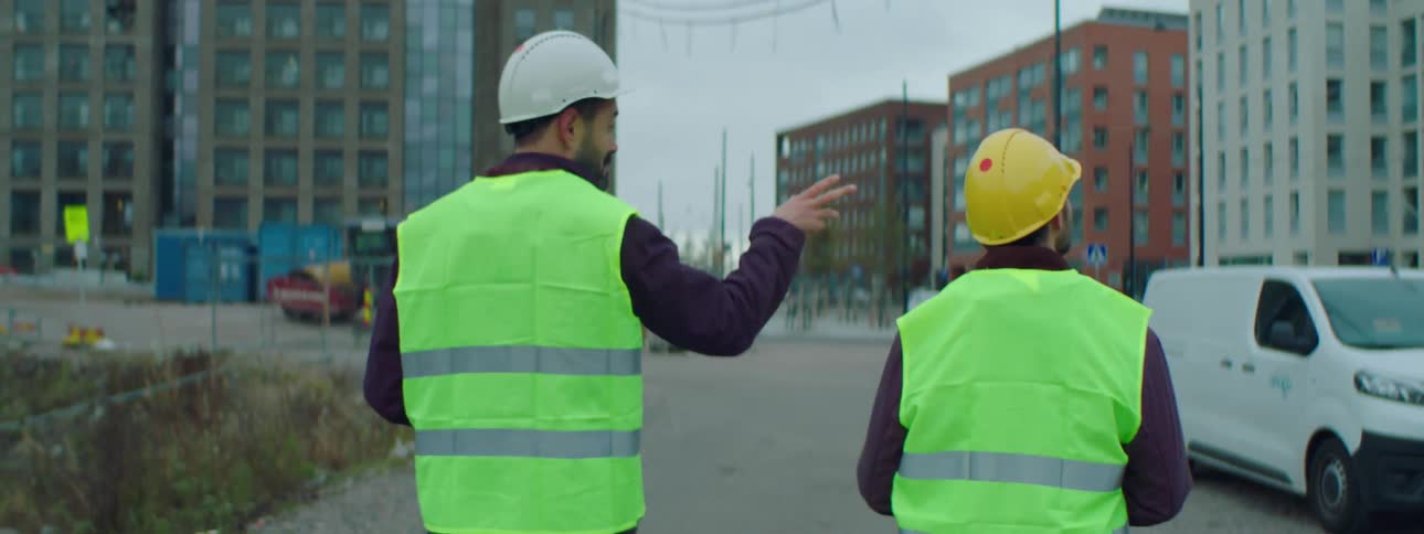 Two construction workers talk as they walk down the street wearing hardhats and safety vests