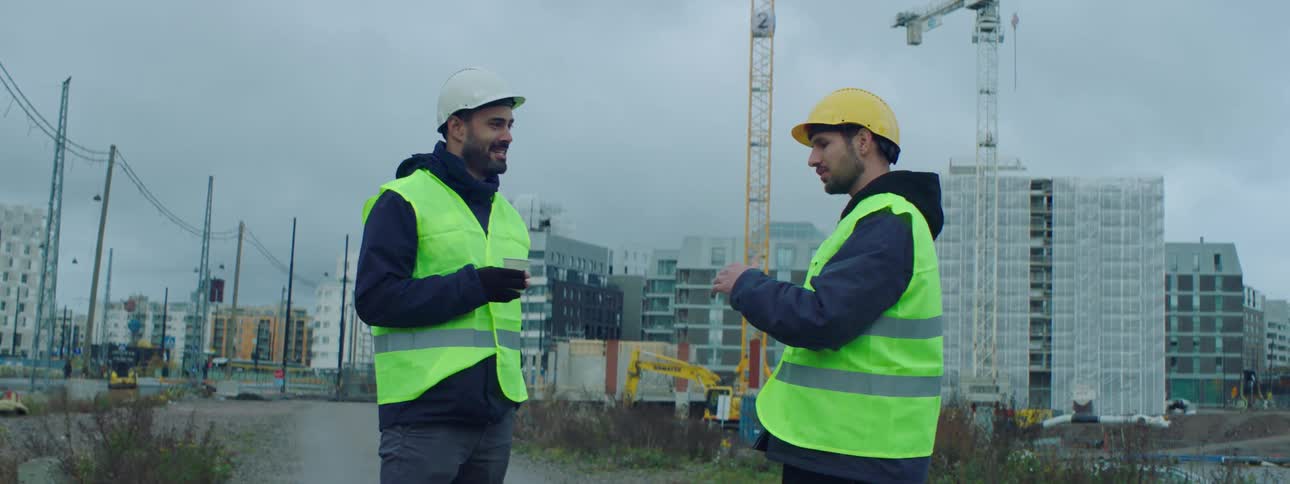 Two male construction workers wearing safety helmets and vests standing at a construction site and talking to each other while one of them holds a cup